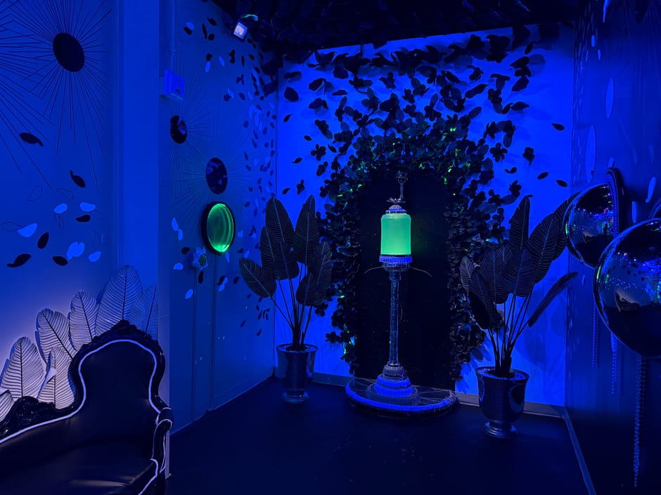 White room with black plants and flowers, glowing in blue light with a green absinthe dispenser
